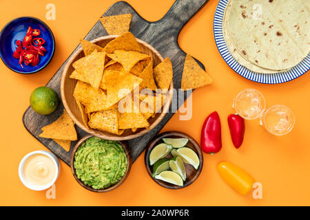 https://l450v.alamy.com/450v/2a0wp6r/mexican-nachos-served-on-wooden-cutting-board-with-guacamole-cheese-sauce-peppers-limes-tequila-and-tortillas-on-orange-background-2a0wp6r.jpg