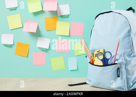 blue backpack with supplies in pocket near multicolored sticky notes on turquoise wall Stock Photo