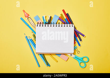 top view of colored pencils, scissors, rubbers and felt pens scattered near blank notebook on yellow Stock Photo