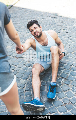 partial view of woman giving hand to injured sportsman sitting on pavement Stock Photo