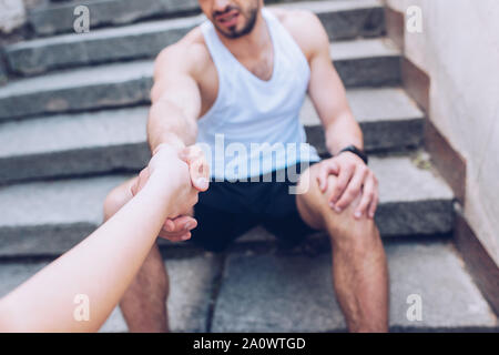 cropped shot of woman giving hand to injured sportsman sitting on stairs and suffering from pain Stock Photo