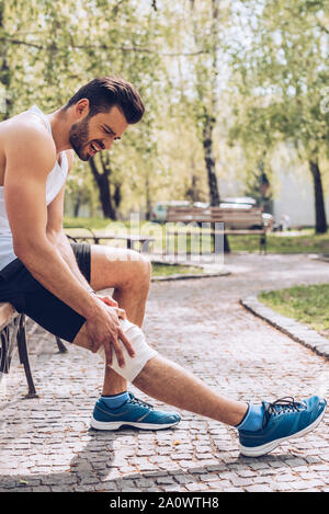 young sportsman suffering from pain while sitting on bench and touching elastic bandage on injured knee Stock Photo