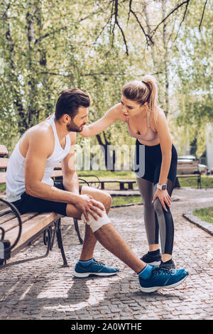 pretty woman in sportswear standing near injured sportsman sitting on bench in park Stock Photo