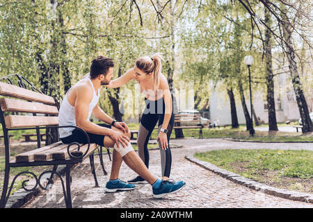 young sportswoman standing near injured sportsman sitting on bench in park Stock Photo