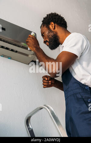 attentive african american repairman standing on ladder and fixing air conditioner Stock Photo