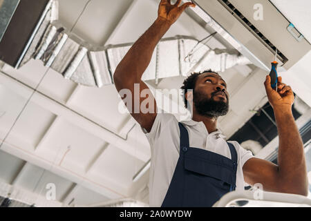 low angle view of african american handyman repairing air conditioner with screwdriver Stock Photo