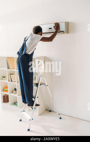 african american handyman in overalls standing on ladder and repairing air conditioner Stock Photo