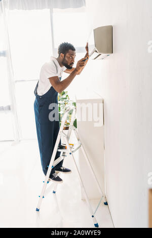 back view of african american repairman standing on ladder and fixing air conditioner Stock Photo