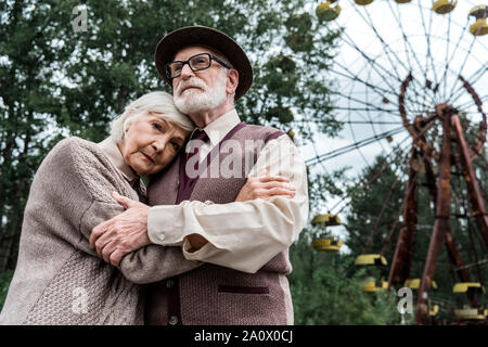bearded senior man hugging with wife near ferris wheel in amusement park Stock Photo