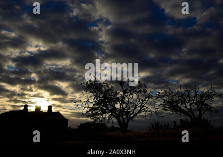 Backlit countryside landscape at sunrise with cloudy sky, house and trees Stock Photo