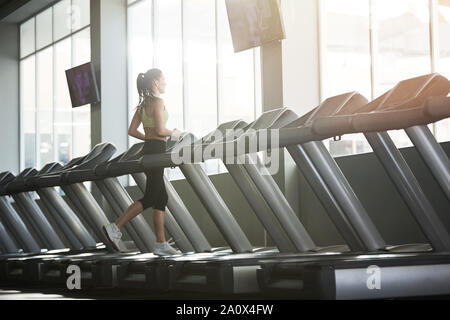 Wide angle back view portrait of young woman running on treadmill alone in empty gym, copy space Stock Photo