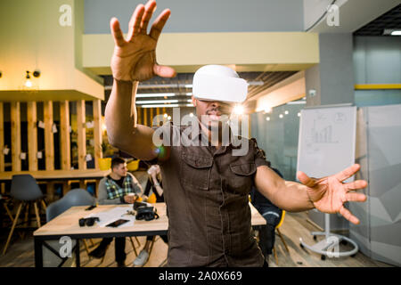 African-American man in casual wear using 3d virtual reality headset. Black gamer wearing goggles, gesturing and having fun in modern coworking place Stock Photo