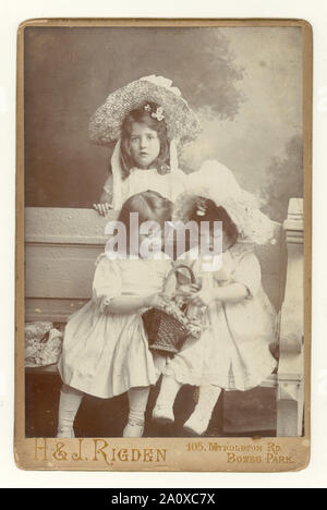 Original antique Edwardian cabinet card, charming studio portrait of three pretty young siblings, sisters, in summer clothes, wearing large straw hats and white smocks, From the studio of Herbert and James Rigden, Bowes Park, London, U.K. circa 1904. Stock Photo