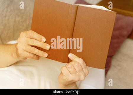 A man is reading a brown cover book while lying on a sofa. Close-up Stock Photo