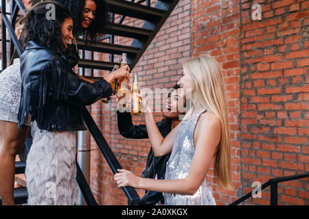 Friends toasting beer bottles on staircase. Stylish women celebrating outdoors with alcohol drinks. Stock Photo