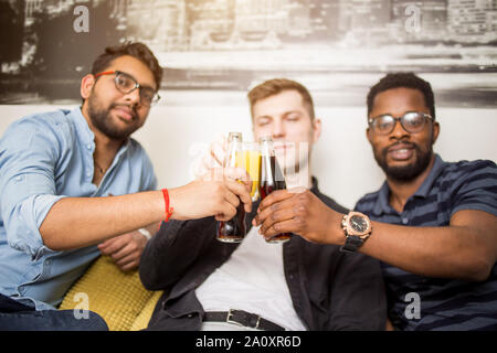 Three handsome multiracial friends in casual clothes sitting on couch clinking bottles of drinks smiling looking at camera. Focus on beverage. People, Stock Photo
