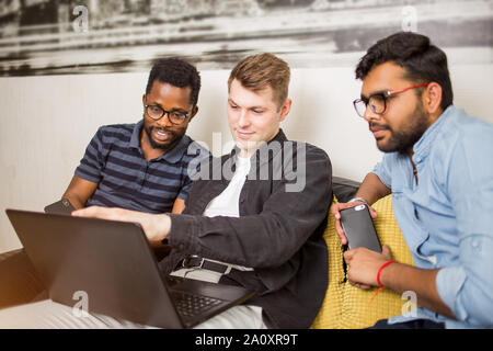 Group of joyful multiethnic students sitting on couch looking at screen of laptop computer, watching film or making presentation togeher. Friends work Stock Photo