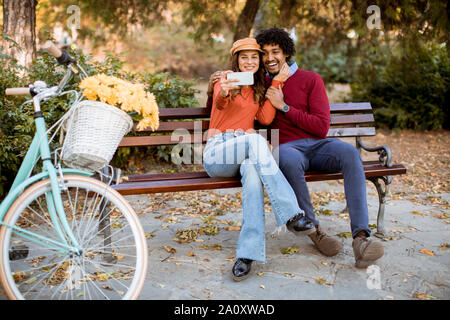 Multiratial young loving couple sitting on bench and taking selfie with mobile phone in the autumn city park Stock Photo