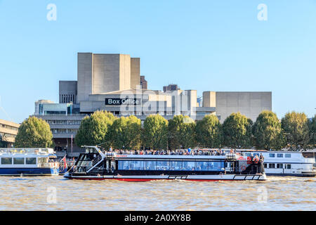 A City Cruises tour boat passing in front of the National Theatre, across the River Thames from the Victoria Embankment, London, UK