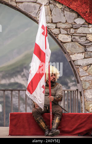 6-16-2019 Military Highway Georgia - Man dressed in traditional Georgian clothing with fur hat holding flag sits in Russia–Georgia Friendship Monument Stock Photo