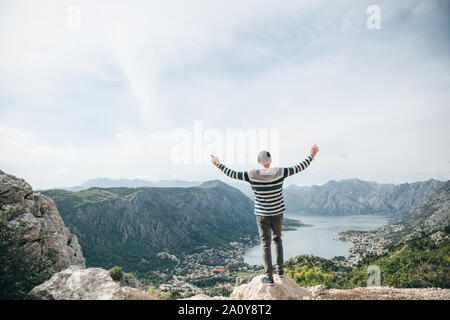 A guy or a tourist admires a beautiful view of the Bay of Kotor in Montenegro. He raised his hands up and shows how happy and free he is. Stock Photo