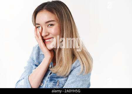 Tenderness, sincere emotions wellbeing concept. Silly feminine asian blond girl touch face shy flirty sensually look camera half-turned smiling giggli Stock Photo