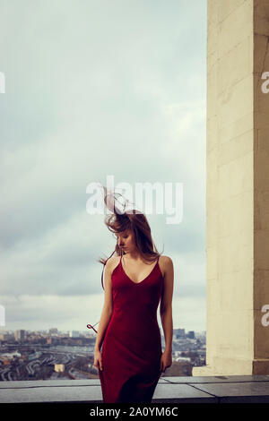 Slim girl in red dress is standing on top of skyscraper, high above the megacity,spreading behind her till the horizon.Her hair is blowing in the wind Stock Photo