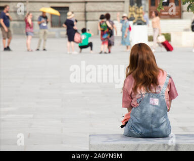 Belgrade, Serbia - September 12, 2019 : One young teenage girl with ginger hair in jeans overall sitting and waiting on city bench on town square, and Stock Photo