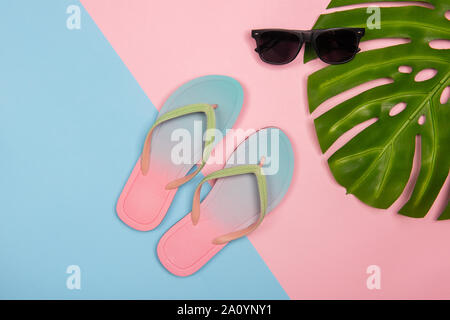 Stylish beach flip-flops on pink and blue pastel background with monstera leaf and sunglasses, top view. Summer minimalistic concept with copy space. Stock Photo
