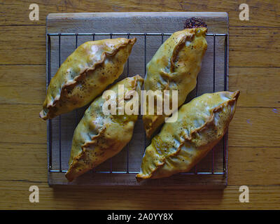 Four Cornish pasties on steel rack on a wooden chopping board, cooling, fresh out of the oven. Stock Photo