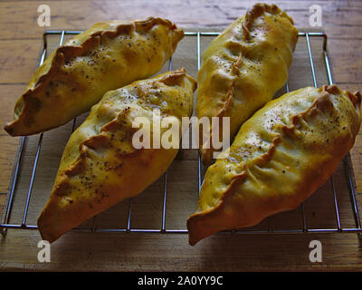 Four Cornish pasties on steel rack on a wooden chopping board, cooling, fresh out of the oven. Stock Photo