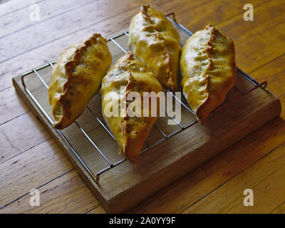 Four Cornish pasties on steel rack on a wooden chopping board, cooling, fresh out of the oven. Stock Photo