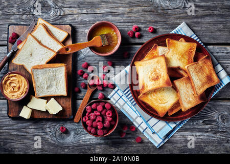 toasts of wheat bread on a plate with melted butter in a bowl and fresh raspberries on a wooden table, ingredients for cooking sandwiches with peanut Stock Photo