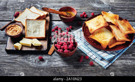 toasts of wheat bread on plate with melted butter in a bowl and fresh raspberries on a wooden table, ingredients for cooking sandwiches with peanut bu Stock Photo