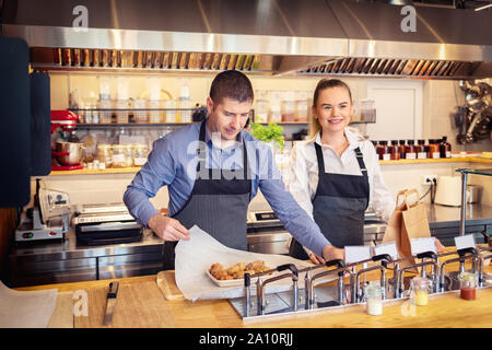 Cheerful waiters wearing apron serving takeaway food at counter in small restaurant Stock Photo