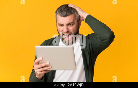 Angry bearded man holding tablet, shouting and touching his head Stock Photo