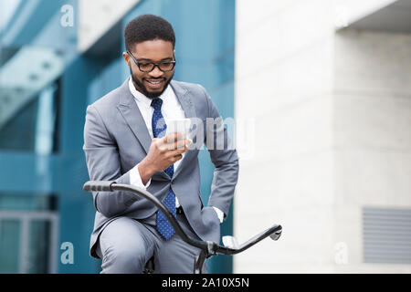 Happy afro businessman using smartphone, riding on bicycle Stock Photo