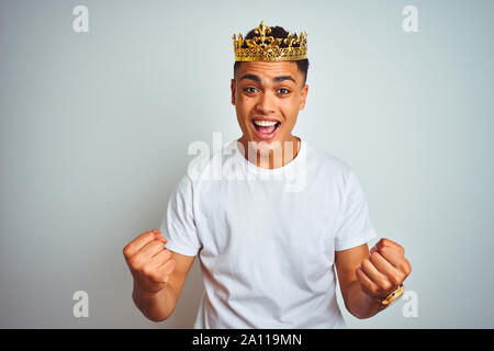 Young brazilian man wearing king crown standing over isolated white background celebrating surprised and amazed for success with arms raised and open Stock Photo