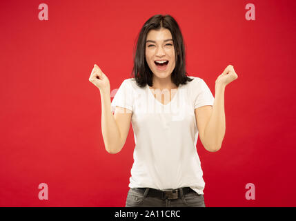 Caucasian young woman's half-length portrait on red studio background. Beautiful female model in white shirt. Concept of human emotions, facial expression. Celebrating, crazy happy, wondered. Stock Photo