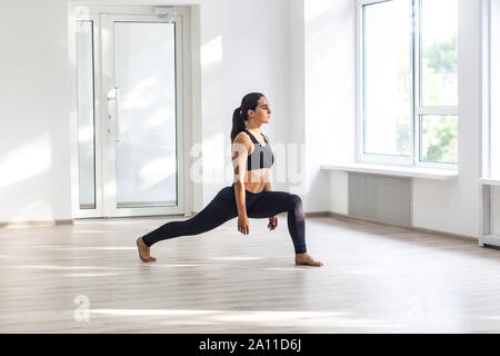 Young attractive sporty woman wearing black sportswear practicing sport exercises, doing lunges near window in gym, stretching legs and standing on as Stock Photo