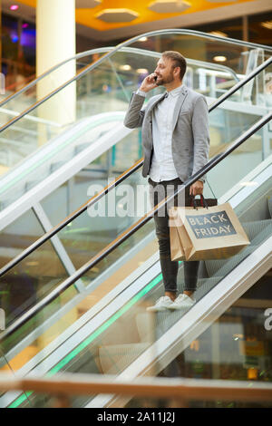 Portrait of fashionable man holding shopping bags with Black Friday inscription while speaking by phone standing on escalator in mall, copy space Stock Photo
