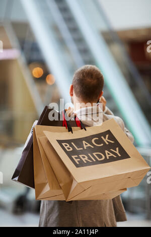 Back view portrait of fashionable man holding shopping bags with Black Friday inscription while speaking by phone standing against escalator in mall, copy space Stock Photo