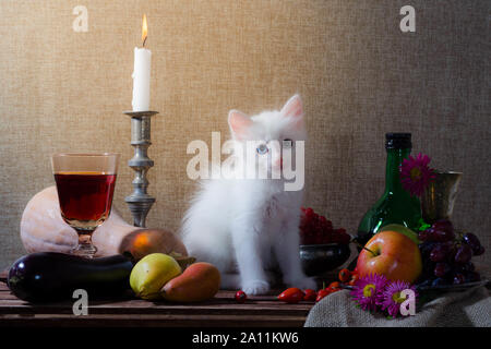 rustic still life with white kitten pumpkin grapes dog rose green bottle grapes viburnum apples glass and tin glasses and a burning candle in a candle Stock Photo