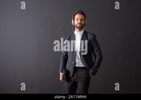 A serious handsome young businessman in a suit holding a tablet and listening to music in front of a dark grey background in a studio. Stock Photo
