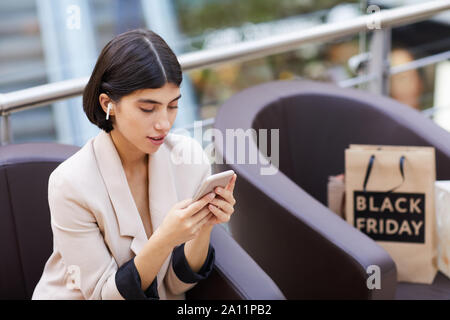 High angle portrait of beautiful young woman wearing wireless earphones using smartphone while taking break from shopping in mall, copy space Stock Photo