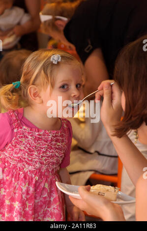 Candid authentic portrait of an adorable cute 3 years of age old blonde hair little girl being fed by her mum at a kid's party. Stock Photo