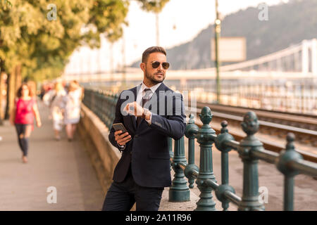 A handsome young businessman standing next to a tramline and using his smartphone on a sunny day. Stock Photo