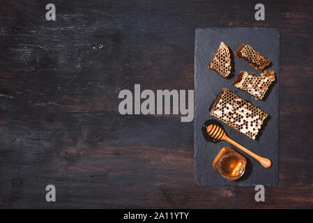 Honeycomb and honey jar with dipper on black slate tray over dark backdrop. Copy space. Top view Stock Photo