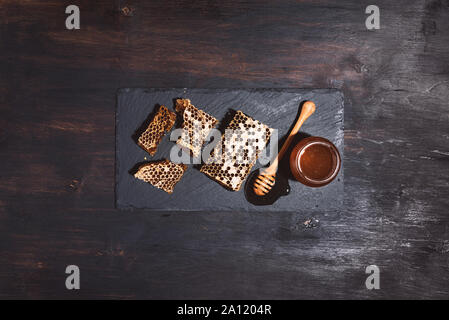 Honeycomb and honey jar with dipper on black slate tray over dark backdrop. Copy space. Top view Stock Photo