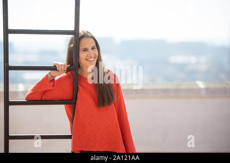Chica adolescente feliz en la azotea mirando a la cámara apoyada en una escalerilla Stock Photo